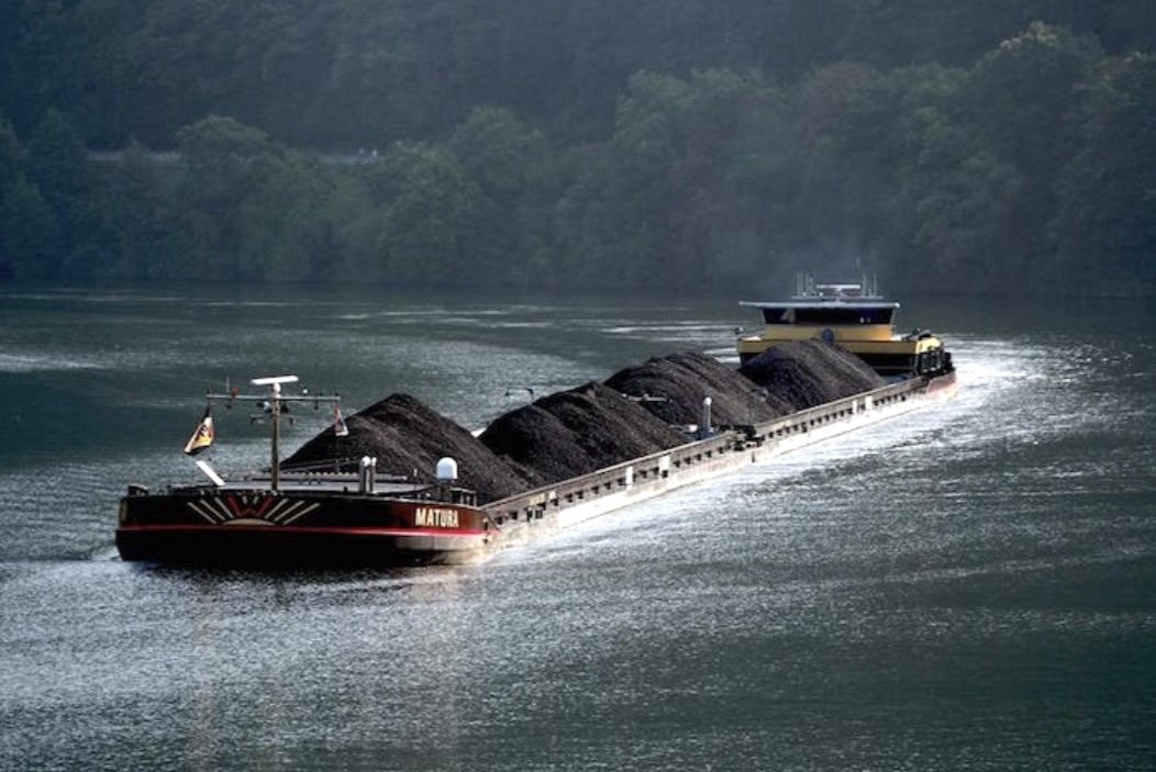 Sebuah kapal tongkang mengangkut batu bara melintasi sungai di tengah kawasan hutan. Impor batu bara global tercatat turun signifikan pada kuartal pertama 2025, seiring lonjakan produksi energi bersih di negara-negara besar. Foto: Construction Week.